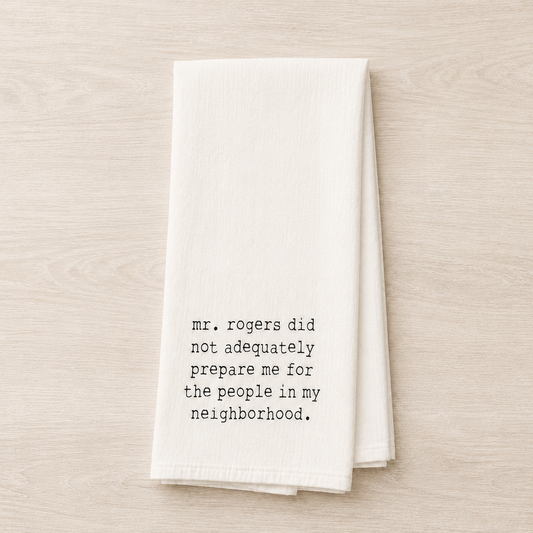 A folded white kitchen towel with black printed text reading 'mr. rogers did not adequately prepare me for the people in my neighborhood.