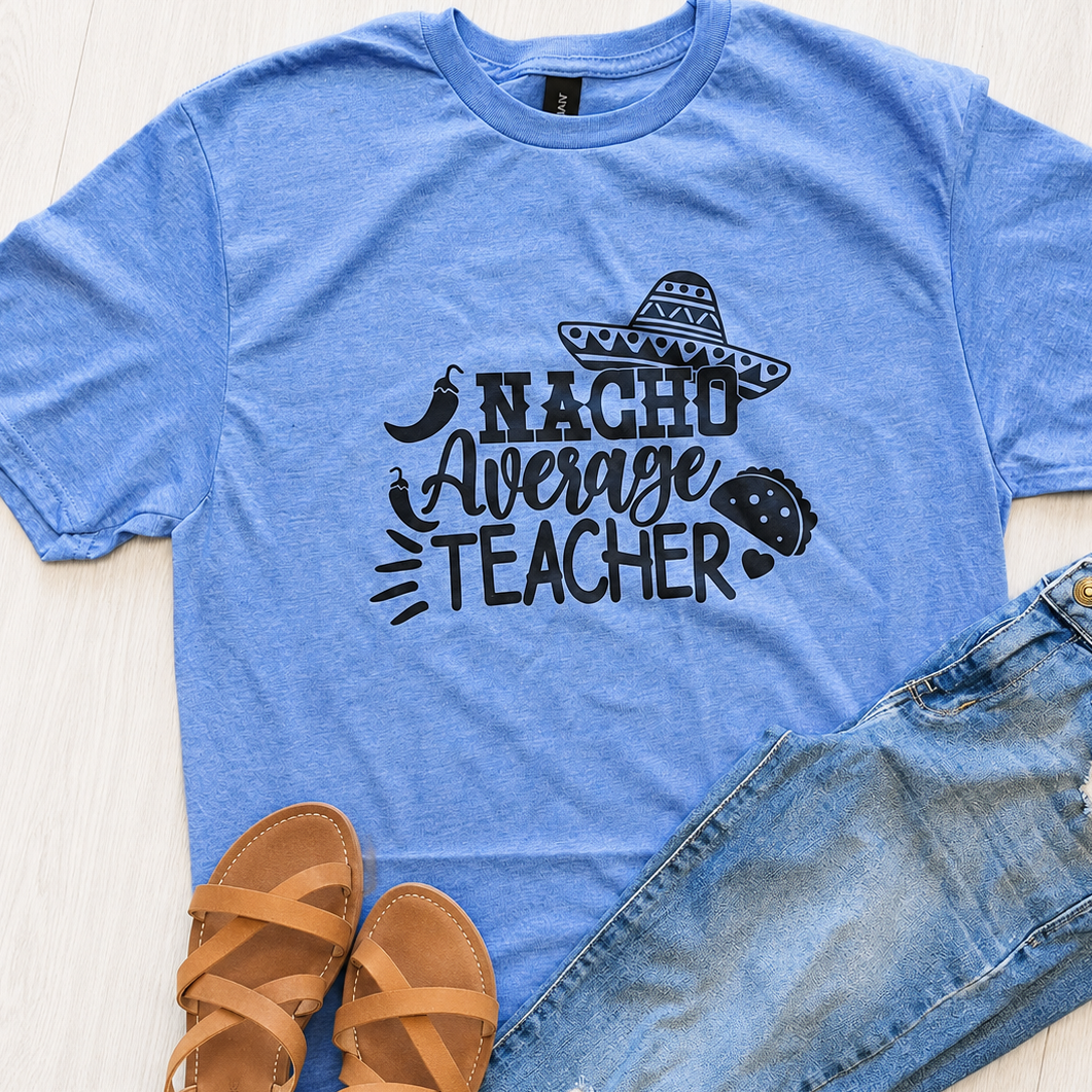 A heather blue graphic t-shirt with a black 'Nacho Average Teacher' design, styled in a flat lay with distressed blue jeans and tan sandals on a light wood floor.