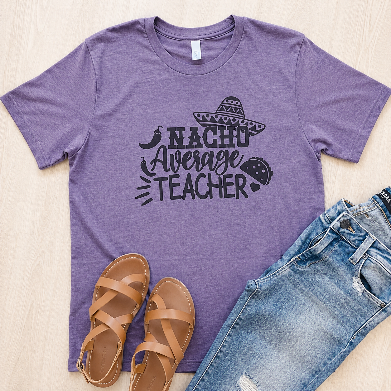 A heather purple short-sleeve graphic T-shirt laid flat on a light wood surface, styled with tan strappy sandals and blue jeans. The shirt has a black printed slogan reading “NACHO Average TEACHER” with small food and hat motifs.