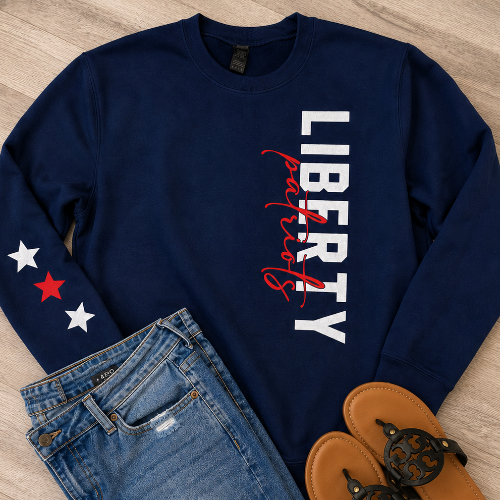 A navy blue long-sleeve sweatshirt with 'LIBERTY' in white block letters and red script text on the front, styled in a flat lay with blue distressed denim shorts and tan slide sandals on a light wood floor.