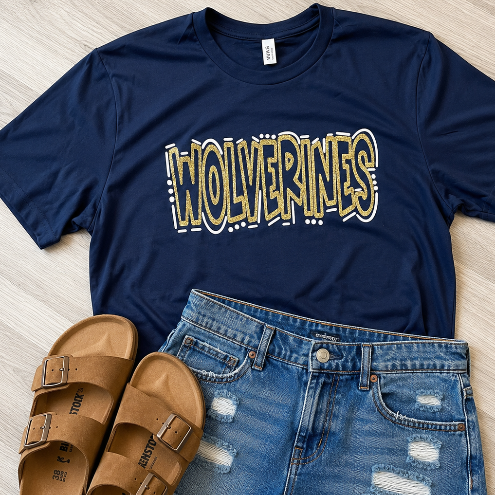 A navy blue graphic t-shirt with 'WOLVERINES' printed in yellow and white, styled in a flat lay with distressed denim shorts and tan slide sandals on a light wood floor.