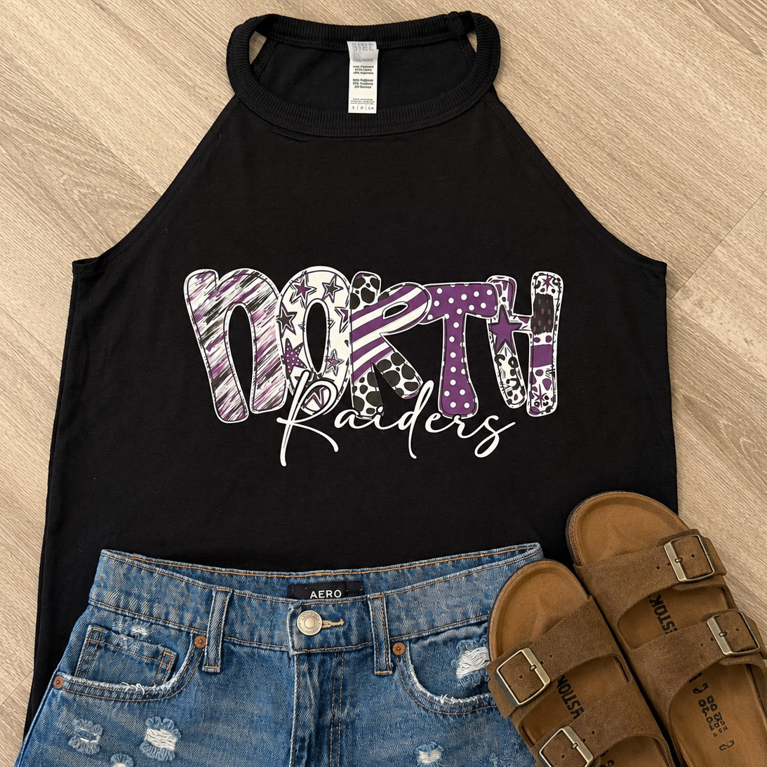 A flat-lay outfit arrangement featuring a black sleeveless tank top with a purple and white patterned 'North Raiders' graphic, paired with distressed blue denim shorts and brown slide sandals on a light wood floor background.