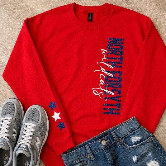 A red crewneck sweatshirt laid flat on a light wood floor, styled with gray New Balance sneakers and distressed blue jeans. The sweatshirt features vertical text reading 'NORTH FORGE' in blue and white script on the right chest, and two stars on the left sleeve in blue and white.