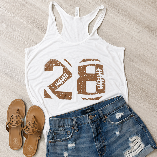 A white racerback tank top with a distressed brown '28 football' graphic, styled in a flat lay with blue distressed denim shorts and tan leather sandals on a light wood floor.