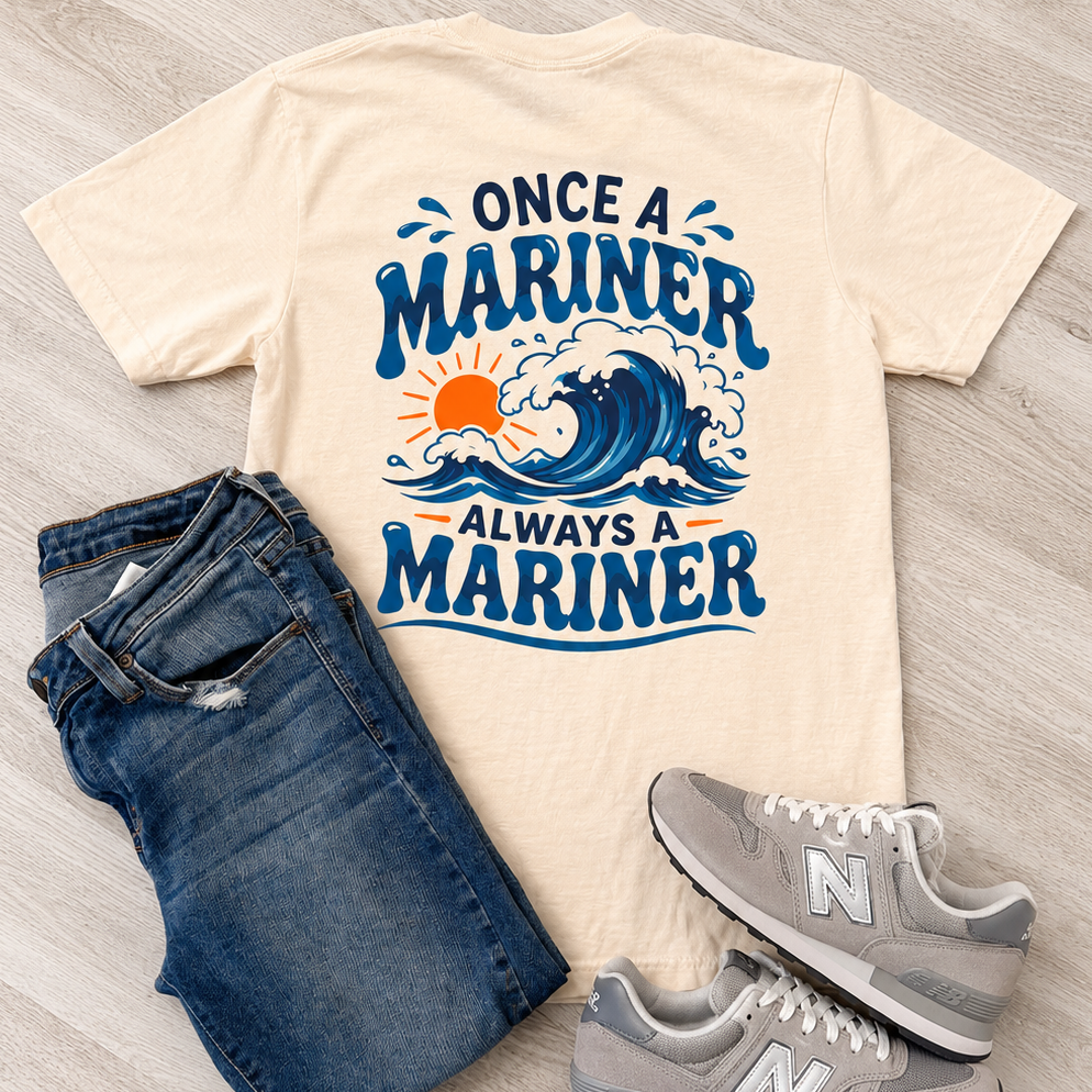 A flat-lay outfit arrangement featuring a cream-colored graphic t-shirt with blue and orange text reading 'ONCE A MARINER ALWAYS A MARINER' over a wave design, styled with blue jeans and gray New Balance sneakers on a light wood floor.