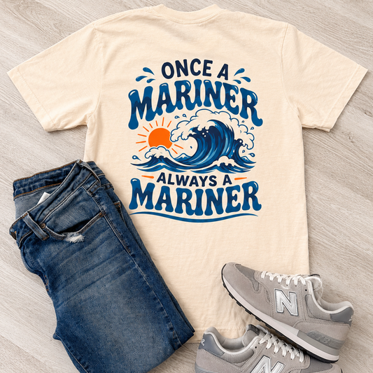 A flat-lay outfit arrangement featuring a cream-colored graphic t-shirt with blue and orange text reading 'ONCE A MARINER ALWAYS A MARINER' over a wave design, styled with blue jeans and gray New Balance sneakers on a light wood floor.