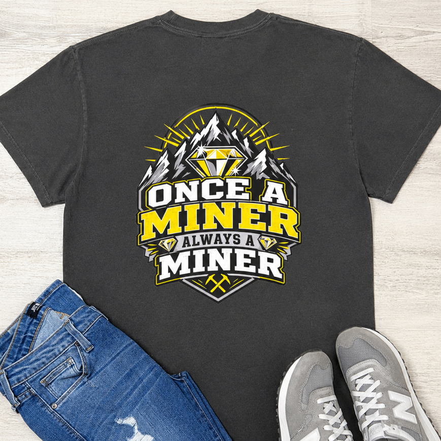 A dark gray graphic t-shirt featuring a 'Once a Miner Always a Miner' design with a gold and white diamond and mountain illustration, styled in a flat lay with blue jeans and gray New Balance sneakers on a light wood background.