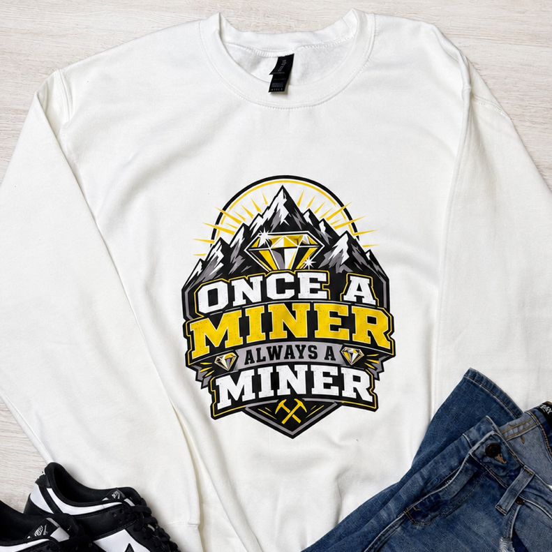 A white crewneck sweatshirt with a 'Once a Miner Always a Miner' graphic, styled in a flat lay with blue jeans and black-and-white sneakers on a light wood floor.