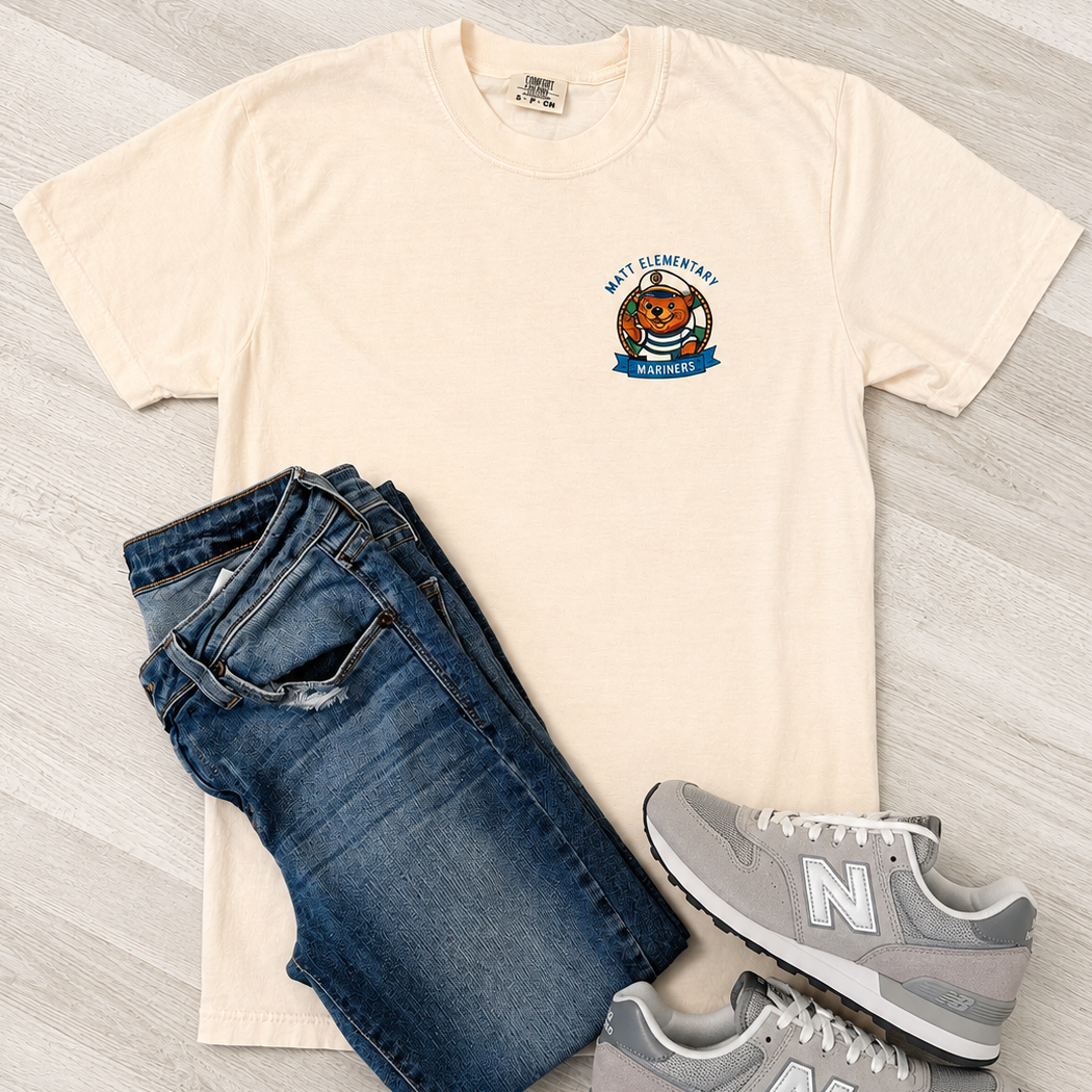 A cream-colored t-shirt with a 'New Orleans Tigers' graphic on the left chest, styled in a flat lay with dark blue jeans and gray New Balance sneakers on a light wood floor.