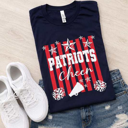A navy blue graphic T-shirt laid flat on a light wood surface, styled with white sneakers and distressed denim shorts. The shirt features red and white vertical stripes, white stars, the word 'PATRIOTS' in white block letters, 'Cheer' in white script, and white pom-pom and megaphone graphics.