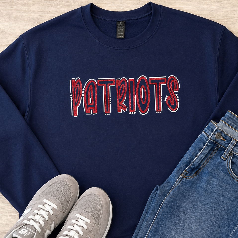 A navy blue crewneck sweatshirt with 'PATRIOTS' printed across the chest in a red, white, and blue flag pattern, styled in a flat lay with blue jeans and gray New Balance sneakers on a light wood background.