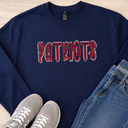 A navy blue crewneck sweatshirt with 'PATRIOTS' printed across the chest in a red, white, and blue flag pattern, styled in a flat lay with blue jeans and gray New Balance sneakers on a light wood background.