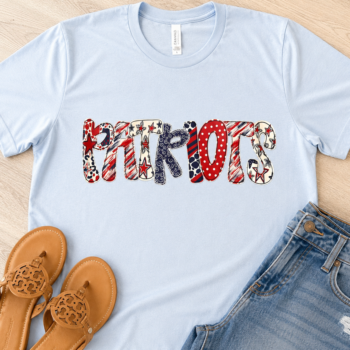 A light blue short-sleeve T-shirt laid flat on a light wood surface, featuring the word "PATRIOTS" across the chest in a patriotic patchwork pattern of red, white, blue, and black. Brown thong sandals and blue jeans are styled around the shirt.