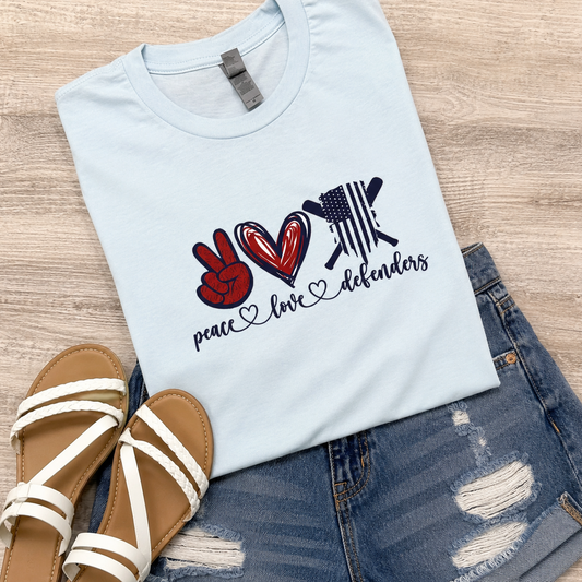 A light blue graphic T-shirt laid flat on a wood surface, styled with distressed denim shorts and white-strap sandals. The shirt graphic shows a peace sign, a red heart, a black-and-white American flag motif, and the words 'peace love defenders' in dark script.