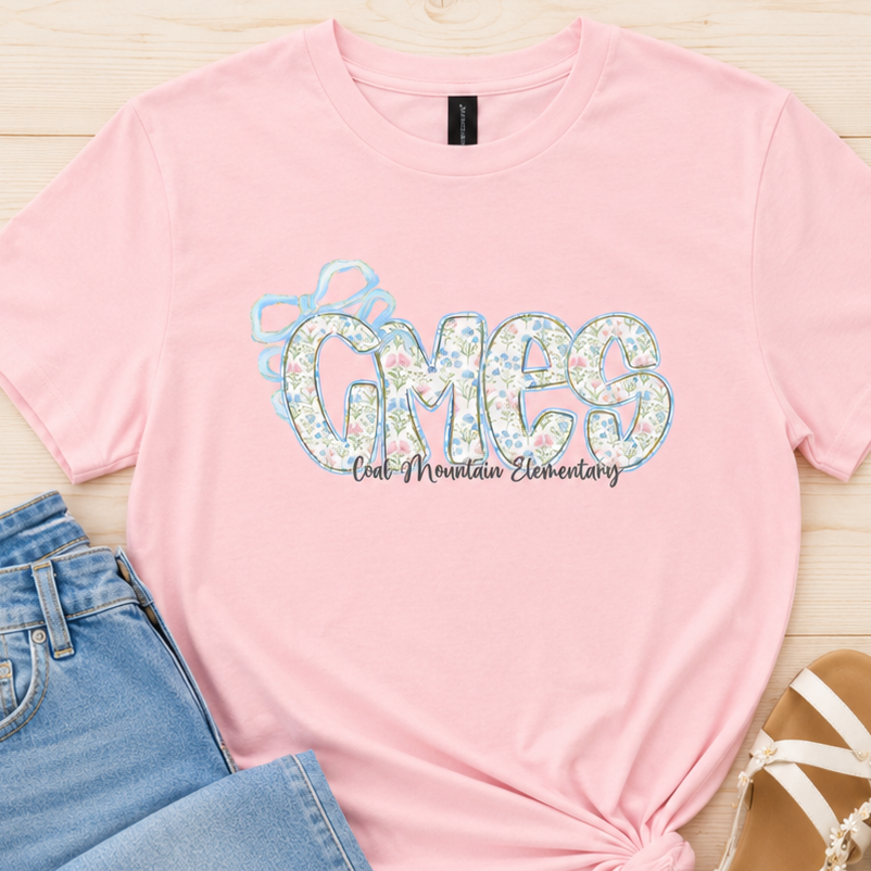 A pink short-sleeve t-shirt with a 'CMES Our Mineral Elementary' graphic, styled in a flat lay with light blue jeans and white sandals on a light wood background.