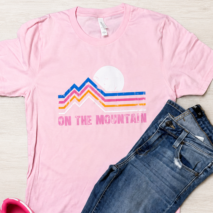 A styled flat-lay outfit featuring a light pink t-shirt with a retro mountain graphic and the text 'ON THE MOUNTAIN', paired with distressed blue jeans and pink and white sneakers on a light wood background.