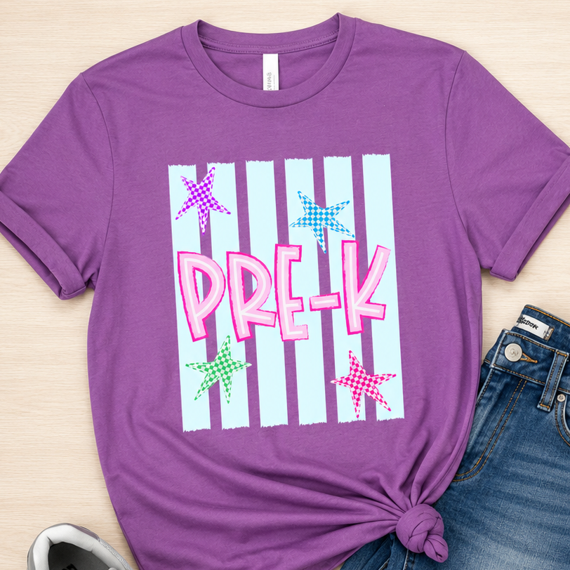 A styled flat-lay featuring a purple short-sleeve graphic T-shirt with 'PREK' in pink over white vertical stripes and colorful star graphics, paired with blue jeans and gray New Balance sneakers on a light wood background.
