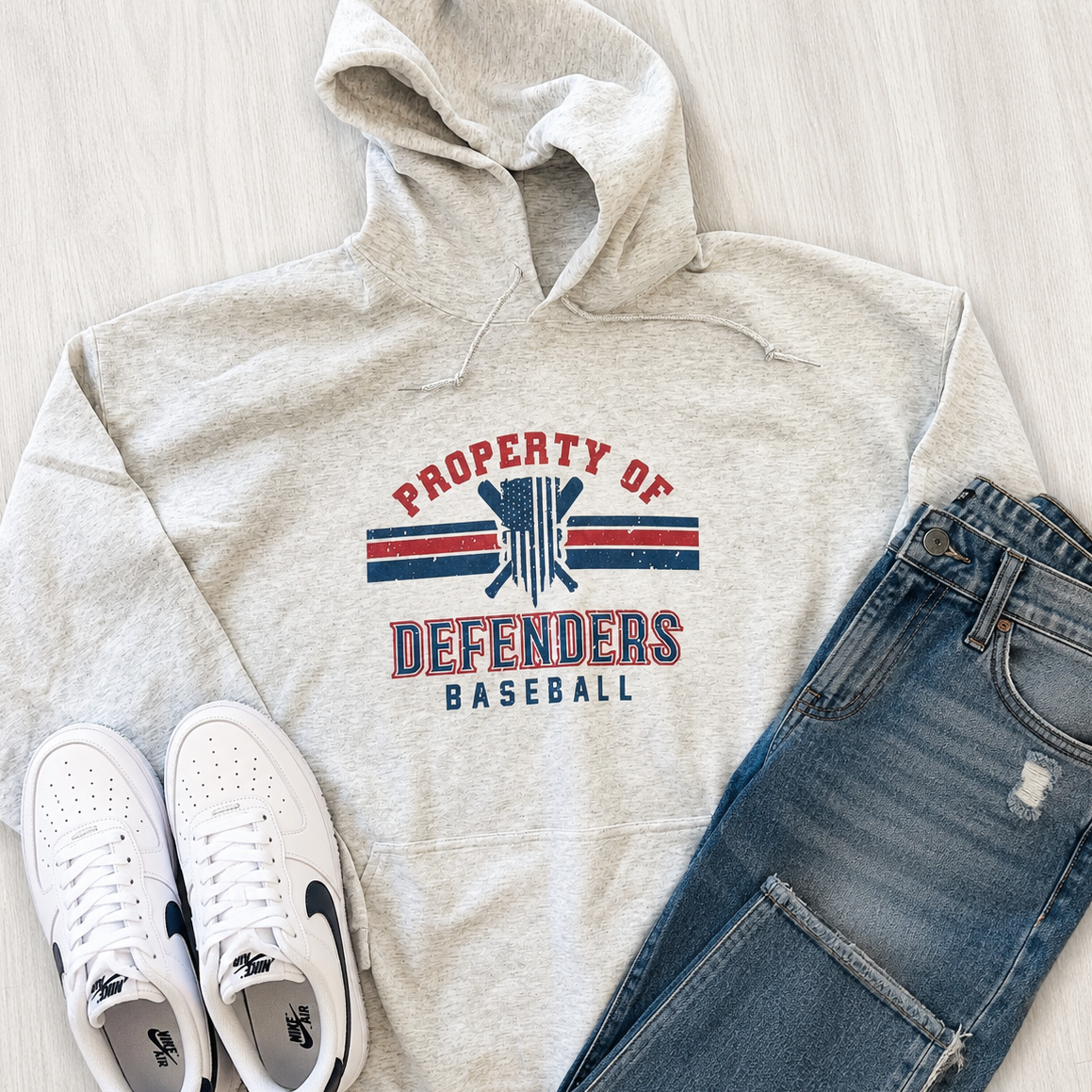 A styled flat-lay of a light heather gray hoodie with a red and blue 'PROPERTY OF DEFENDERS BASEBALL' graphic, paired with blue jeans and white sneakers on a light wood floor.