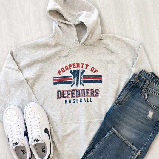 A styled flat-lay of a light heather gray hoodie with a red and blue 'PROPERTY OF DEFENDERS BASEBALL' graphic, paired with blue jeans and white sneakers on a light wood floor.