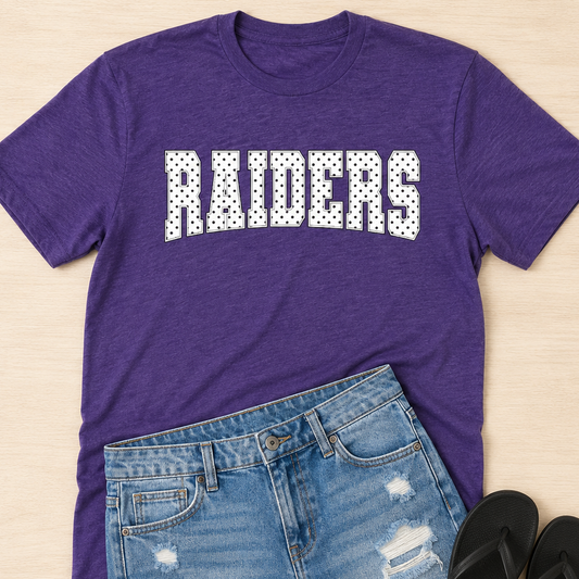 A purple heathered short-sleeve t-shirt with 'RAIDERS' printed across the chest in white block letters with a black polka-dot fill, styled in a flat lay with distressed denim shorts and black flip-flops on a light wood background.