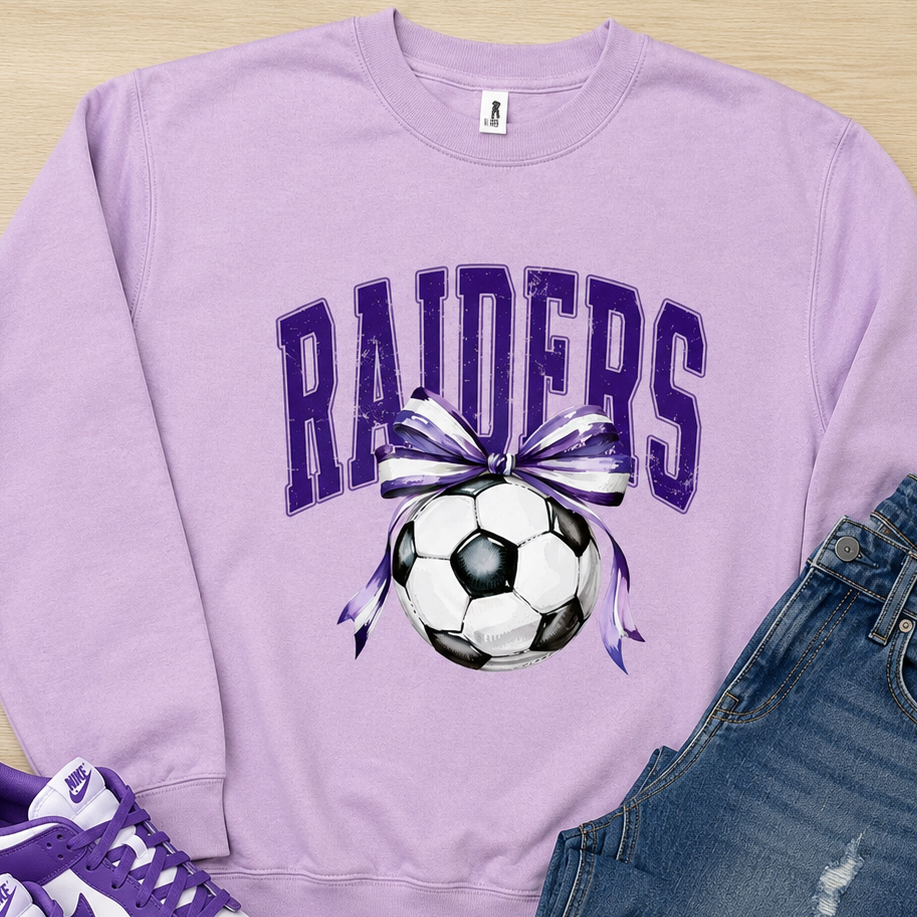 A flat-lay outfit arrangement featuring a light purple sweatshirt with a 'RAIDERS' soccer graphic, paired with blue jeans and purple-and-white sneakers on a light wood background.