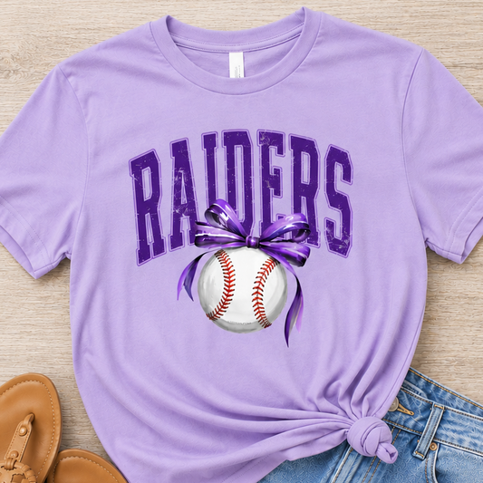 A lavender purple graphic t-shirt featuring the word 'RAIDERS' in purple varsity letters above a baseball with a purple ribbon, styled in a flat lay with denim shorts and tan sandals on a light wood background.