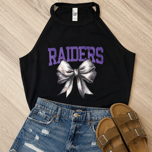 A black halter crop top with purple 'RAIDERS' text and a silver bow graphic, styled in a flat lay with blue denim shorts and tan slide sandals on a light wood floor.