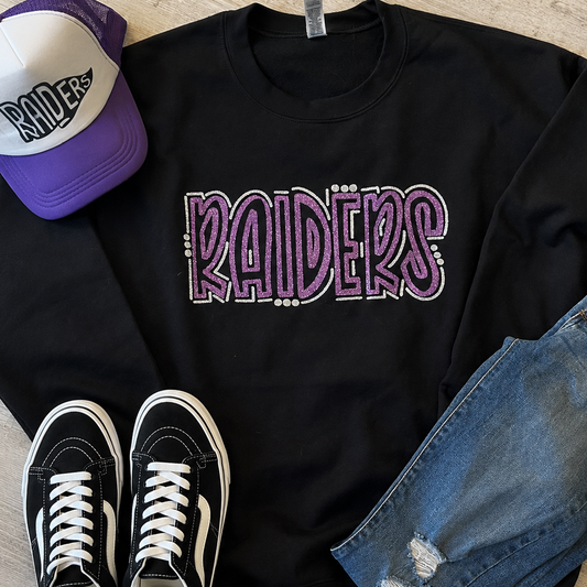 A black crewneck sweatshirt with 'RAIDERS' printed across the chest in purple and white, styled in a flat lay with a purple and white Raiders baseball cap, black sneakers, and blue jeans on a light wood floor.