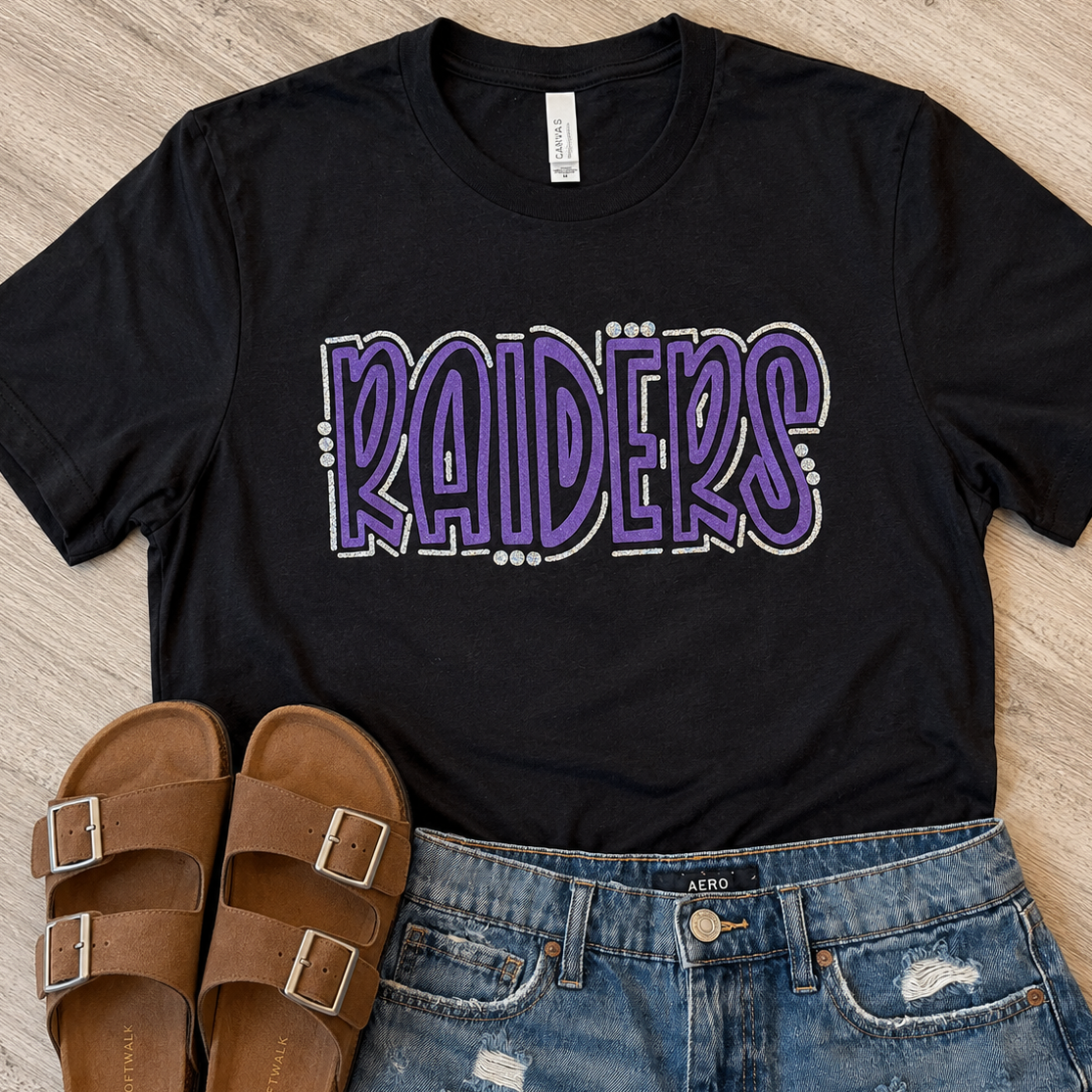 A black short-sleeve graphic T-shirt with the word 'RAIDERS' in large purple outlined lettering, styled flat on a light wood floor with brown double-strap sandals and distressed blue jeans.