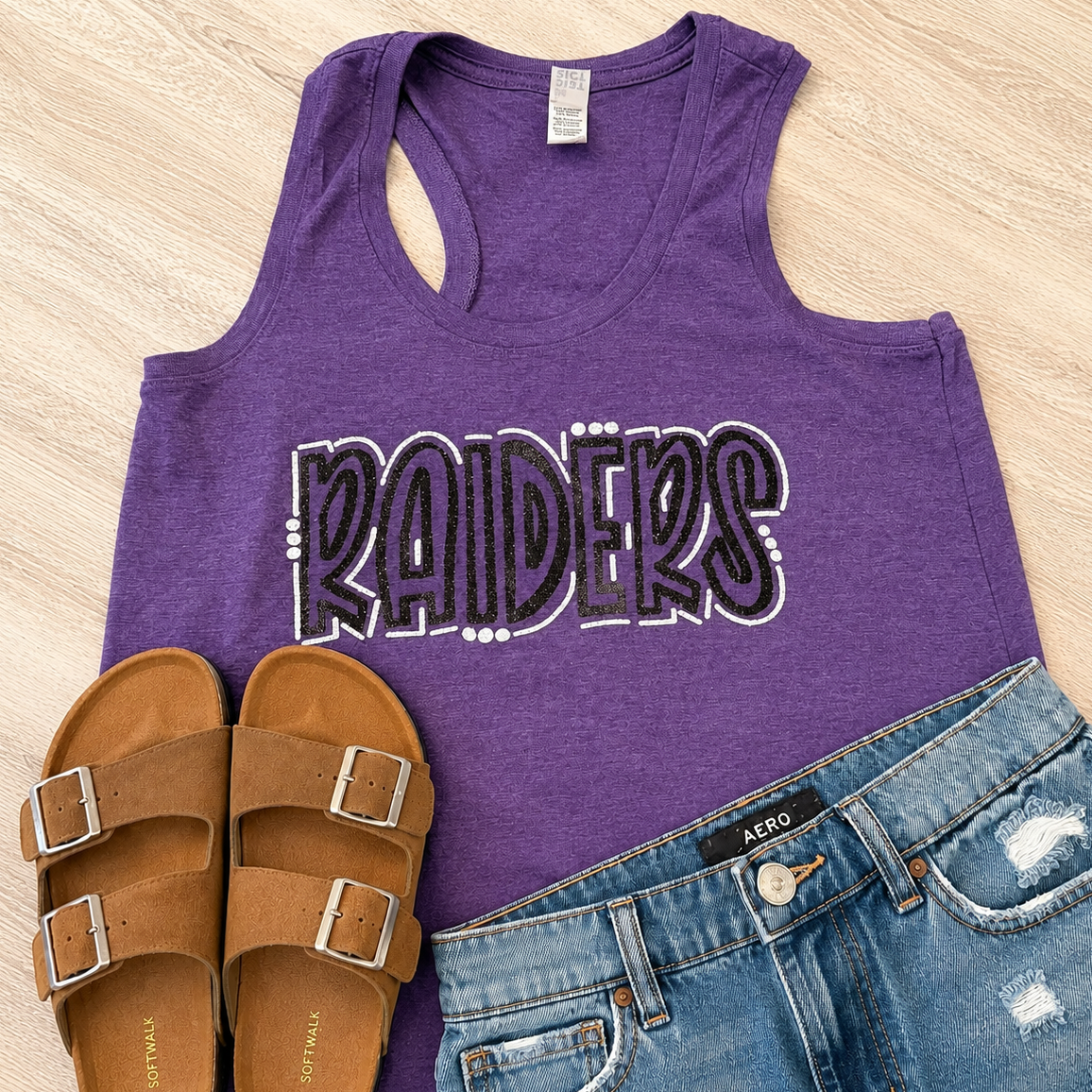 A purple racerback tank top with 'RAIDERS' printed across the front, styled in a flat lay with brown slide sandals and blue distressed denim shorts on a light wood background.