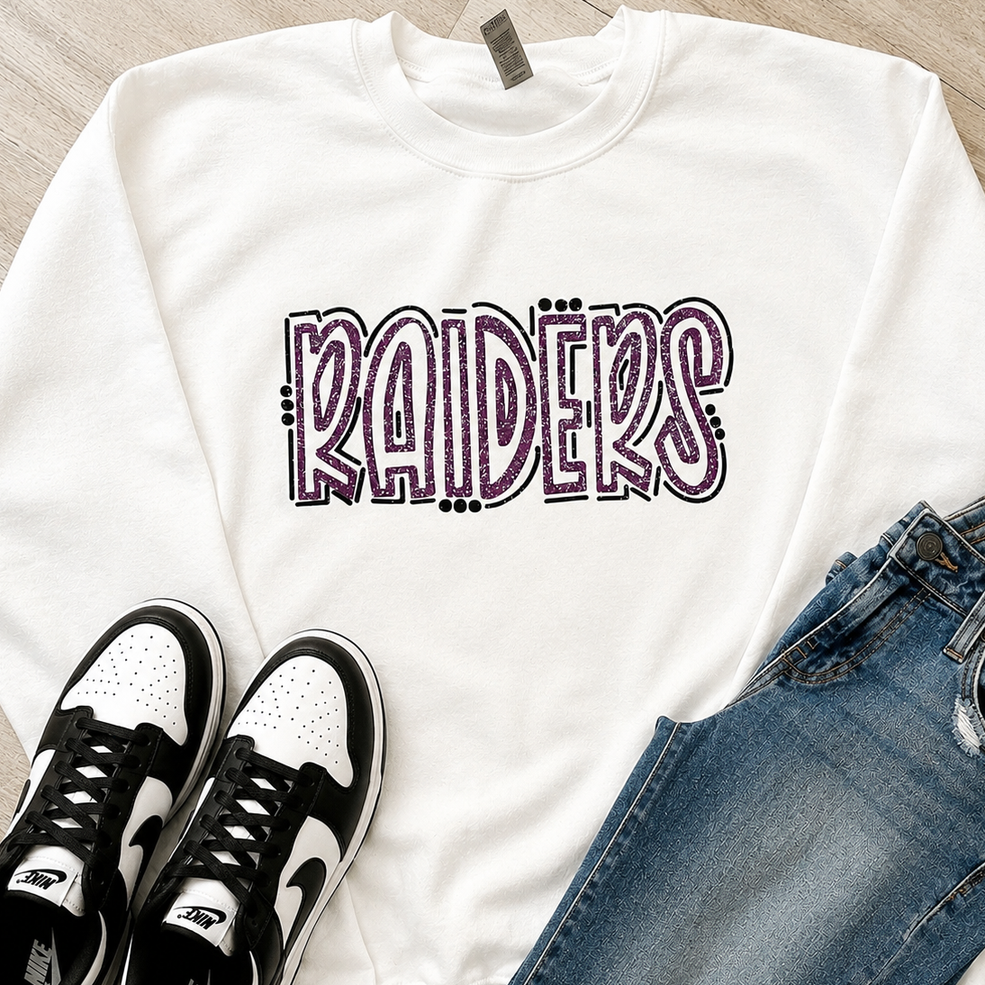 A white crewneck sweatshirt with 'RAIDERS' printed across the chest in a stylized purple and black outline font, styled in a flat lay with blue jeans and black-and-white sneakers on a light wood floor.
