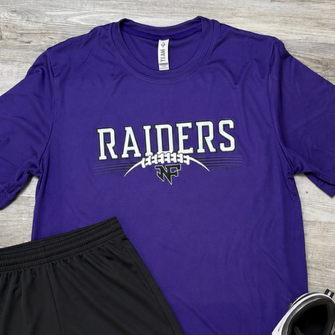 A purple short-sleeve t-shirt with 'RAIDERS' in white text and a small mascot graphic, laid flat on a gray wood-look floor alongside black athletic shorts and black-and-white sneakers.