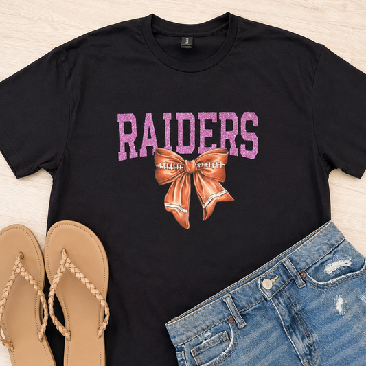 A black short-sleeve graphic T-shirt laid flat on a light wood surface, styled with tan flip-flops and distressed blue denim shorts. The shirt has pink 'RAIDERS' lettering and an orange-and-white bow graphic on the front.
