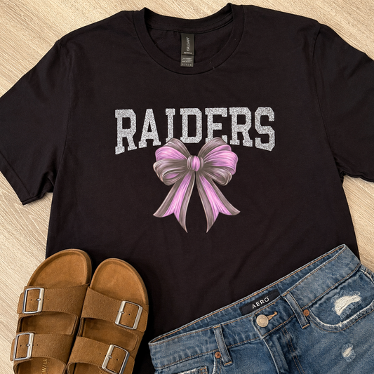 A black graphic t-shirt featuring the word 'RAIDERS' in distressed white letters and a pink and gray bow illustration, styled in a flat lay with brown slide sandals and distressed blue jeans on a light wood background.