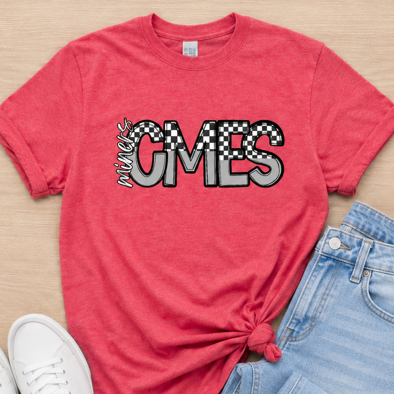 A coral pink graphic t-shirt with 'CMES' in a checkered and zebra-print design, styled in a flat lay with light blue jeans and white sneakers on a light wood background.
