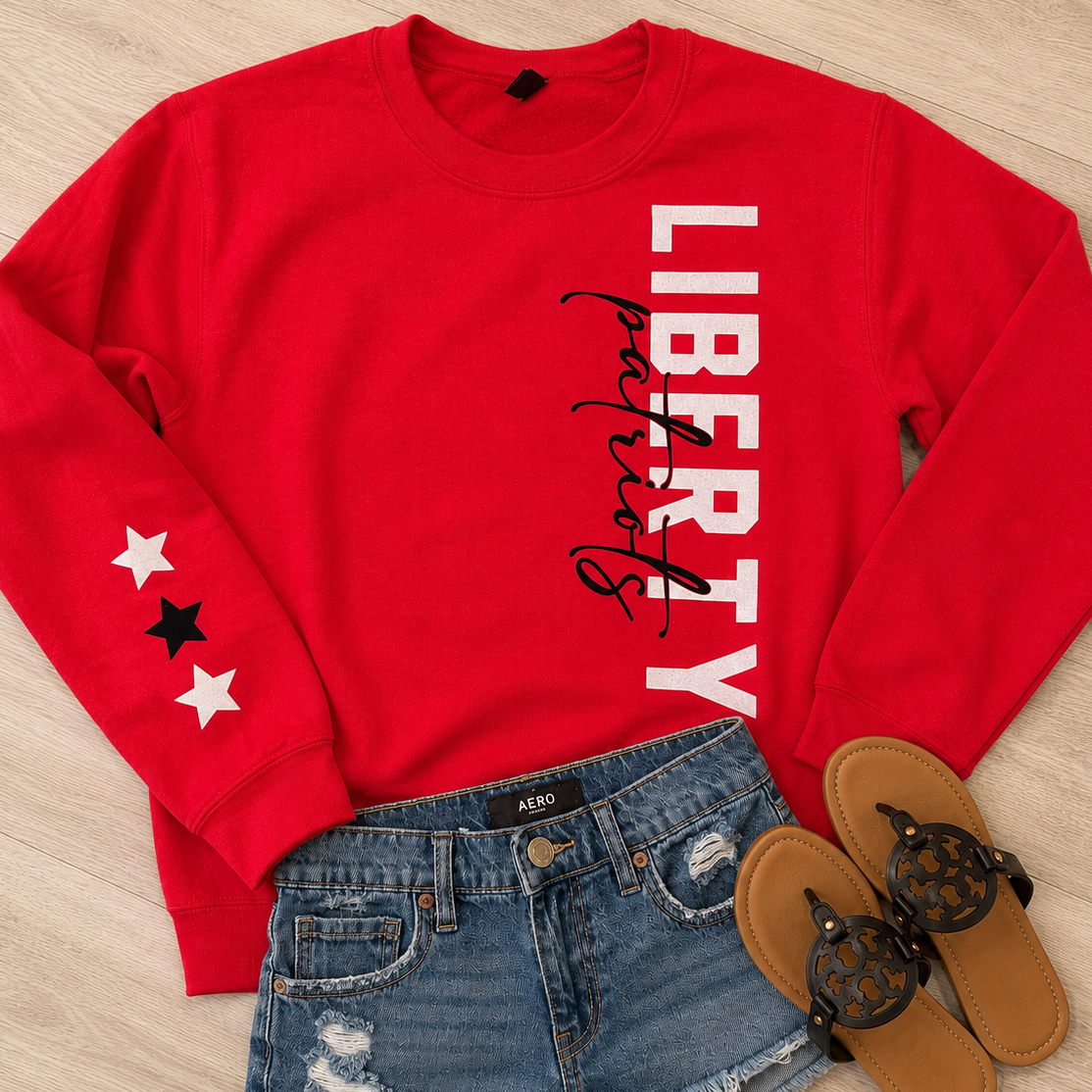A red crewneck sweatshirt with 'LIBERTY' printed vertically in white and black script, styled in a flat lay with blue denim shorts and black sandals on a light wood floor.