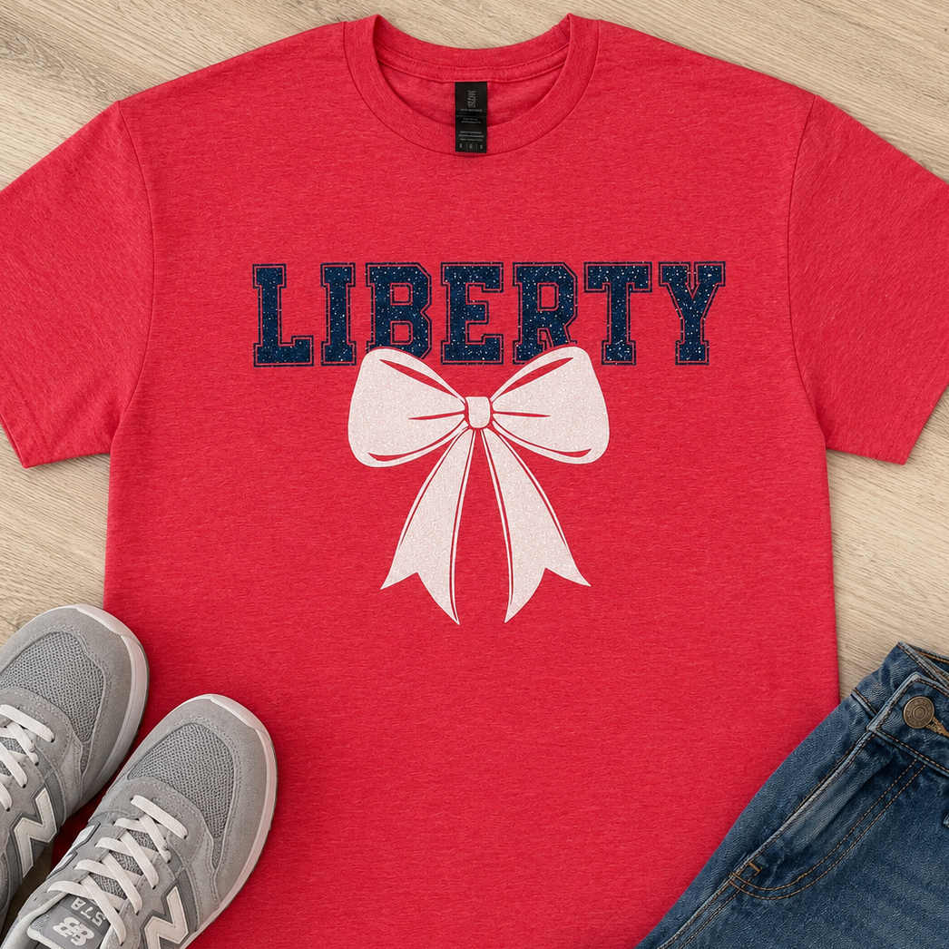 A red short-sleeve T-shirt laid flat on a light wood surface, printed with the word "LIBERTY" in dark navy letters and a large pale pink bow graphic. Gray New Balance sneakers and blue jeans are styled around it as props.