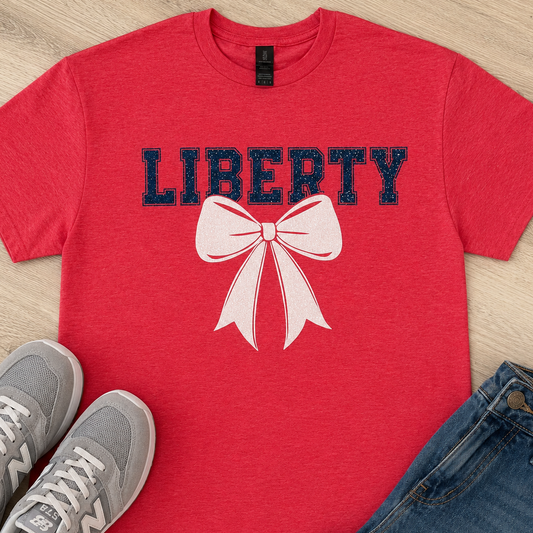 A red short-sleeve T-shirt laid flat on a light wood surface, printed with the word "LIBERTY" in dark navy letters and a large pale pink bow graphic. Gray New Balance sneakers and blue jeans are styled around it as props.