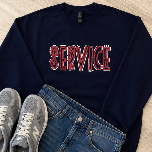 A navy blue crewneck sweatshirt with 'SERVICE' spelled out in red and white rhinestone-style letters, styled in a flat lay with blue jeans and gray New Balance sneakers on a light wood floor.