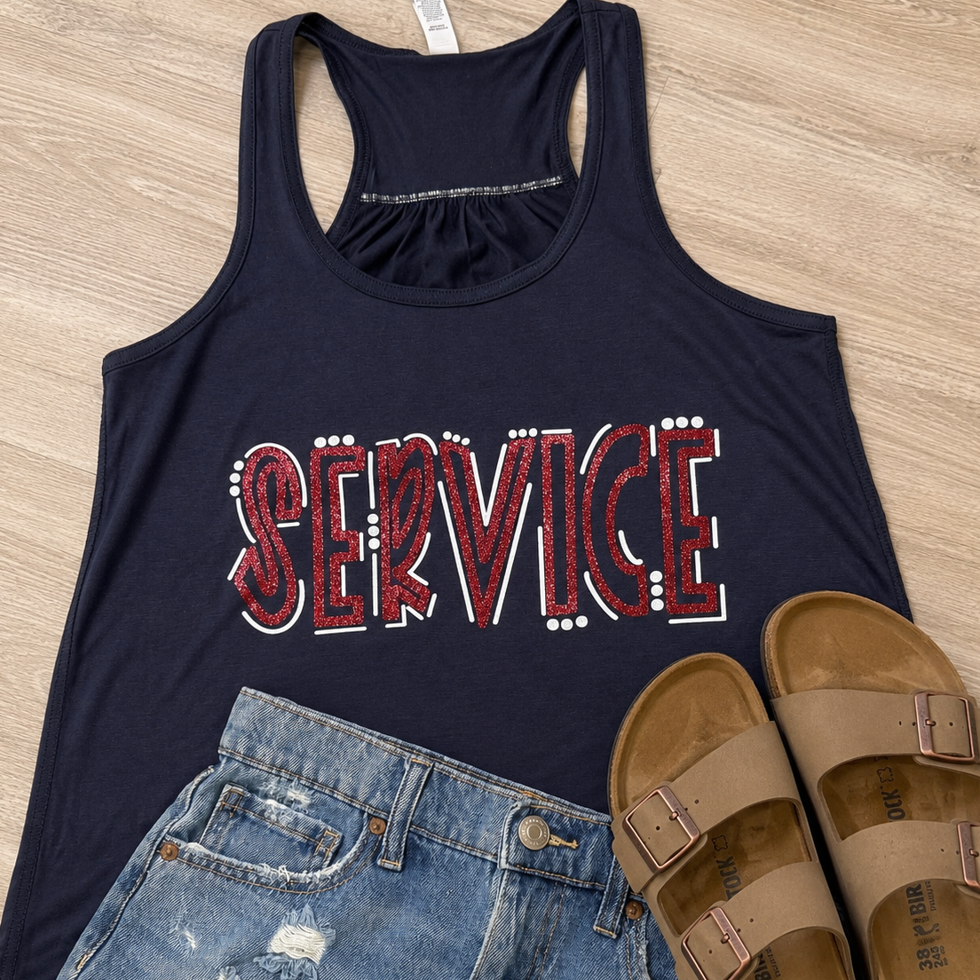 A navy blue racerback tank top with 'SERVICE' printed across the front in red and white, styled in a flat lay with distressed blue denim shorts and tan slide sandals on a light wood floor.
