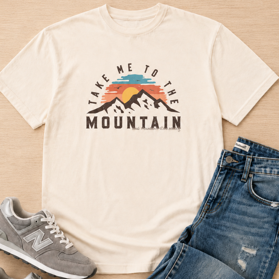 A cream-colored graphic t-shirt with a mountain sunset print reading 'TAKE ME TO THE MOUNTAIN', styled in a flat lay with blue jeans and gray New Balance sneakers on a light wood background.