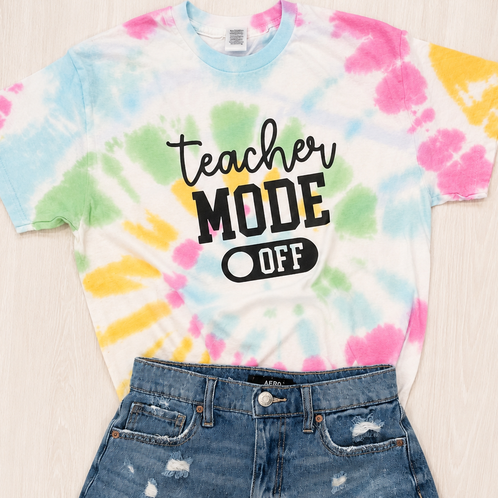 A flat-lay of a white tie-dye t-shirt with 'teacher MODE OFF' printed on the front, styled with distressed denim shorts and brown sandals on a light wood background.