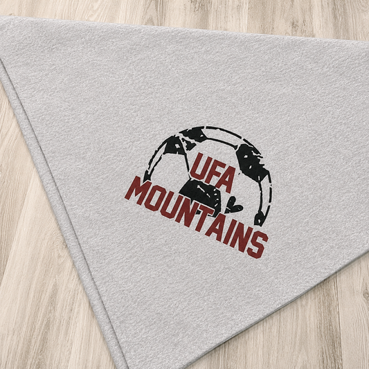 A light gray triangular bandana featuring a black and white soccer ball graphic with the text 'UFA MOUNTAINS' in dark red, laid flat on a light wood floor.