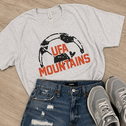 A styled flat-lay outfit featuring a light heather gray graphic t-shirt with 'UFA MOUNTAINS' in orange and black, paired with distressed blue denim shorts and gray New Balance sneakers on a light wood floor.