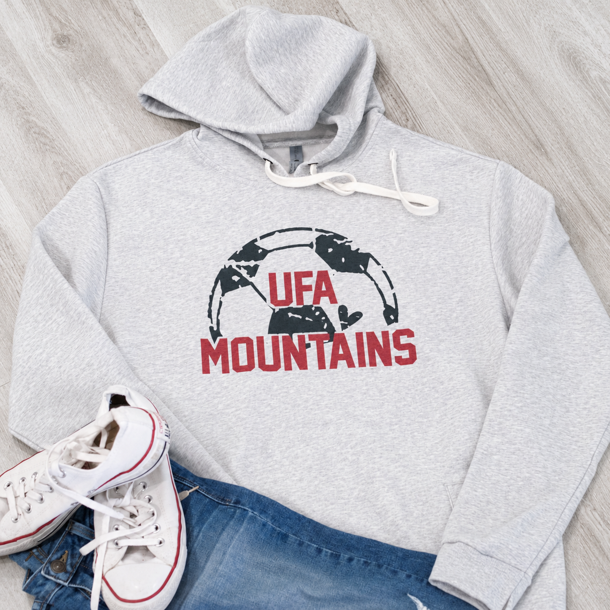 A light heather gray pullover hoodie with a 'UFA MOUNTAINS' graphic in red and black, styled in a flat lay with white sneakers and blue jeans on a light wood floor.