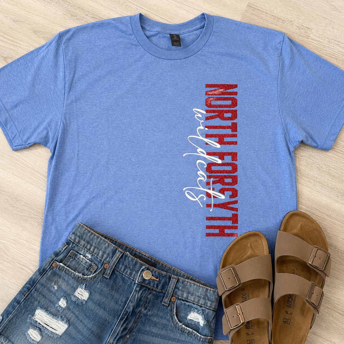 A heather blue short-sleeve T-shirt laid flat on a light wood floor, featuring vertical red and white 'NORTH FORGOTTEN' text on the front. Blue jeans and tan slide sandals are styled around it as a flat-lay outfit.