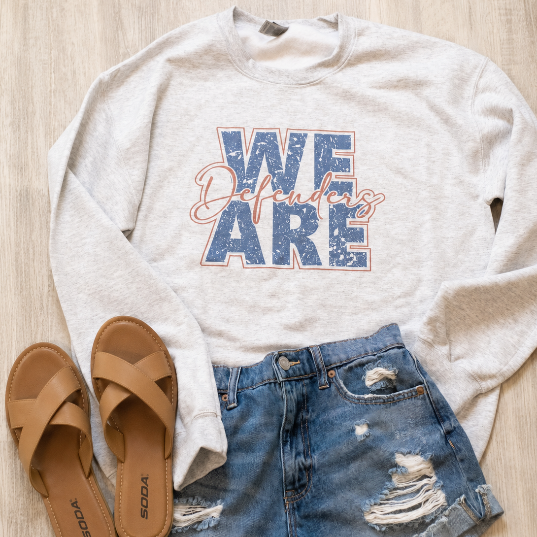 A styled flat-lay featuring a light heather gray sweatshirt with a distressed blue and pink graphic that reads 'WE Defenders', paired with distressed denim shorts and tan sandals on a light wood background.
