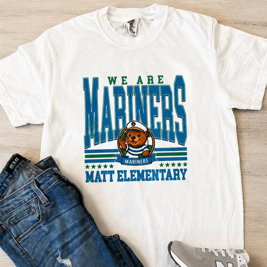 A white graphic t-shirt featuring the text 'WE ARE MARINERS MATT ELEMENTARY' in blue and green, with a cartoon bear mascot wearing a helmet. The shirt is styled in a flat lay with blue jeans and a gray sneaker on a light wood background.