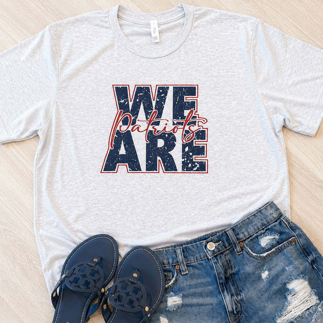 A light heather gray graphic T-shirt laid flat on a light wood surface, styled with distressed blue denim shorts and navy blue Mary Jane sandals. The shirt graphic reads “WE ARE Patriots” in navy with red script and red outline.