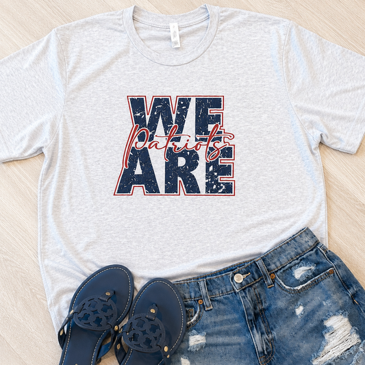 A light heather gray graphic T-shirt laid flat on a light wood surface, styled with distressed blue denim shorts and navy blue Mary Jane sandals. The shirt graphic reads “WE ARE Patriots” in navy with red script and red outline.